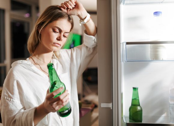 Young woman standing on kitchen at night near opened fridge thoughtfully looking on beer